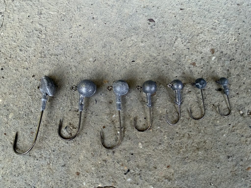 Seven fishing jig heads of different sizes arranged from largest to smallest on a concrete surface
