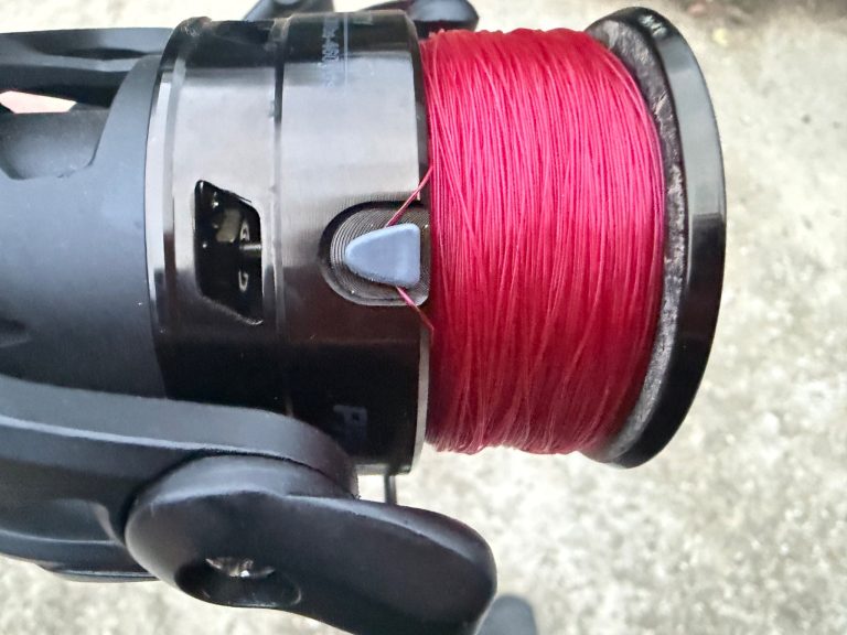 Fishing reel line clip replacement reference showing a gray line clip on a spinning reel spool with red fishing line